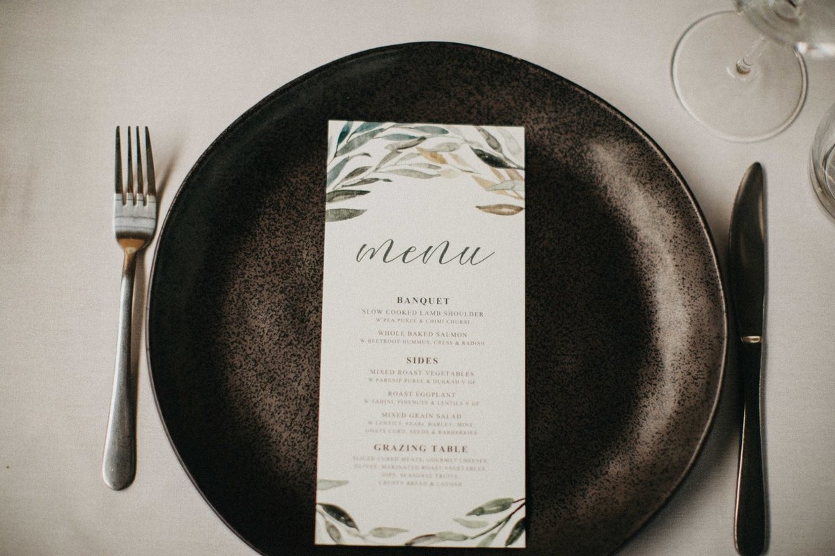 A printed menu is centered on a dark plate beside cutlery. Photograph by documentary wedding photographer Joshua Mikhaiel.
