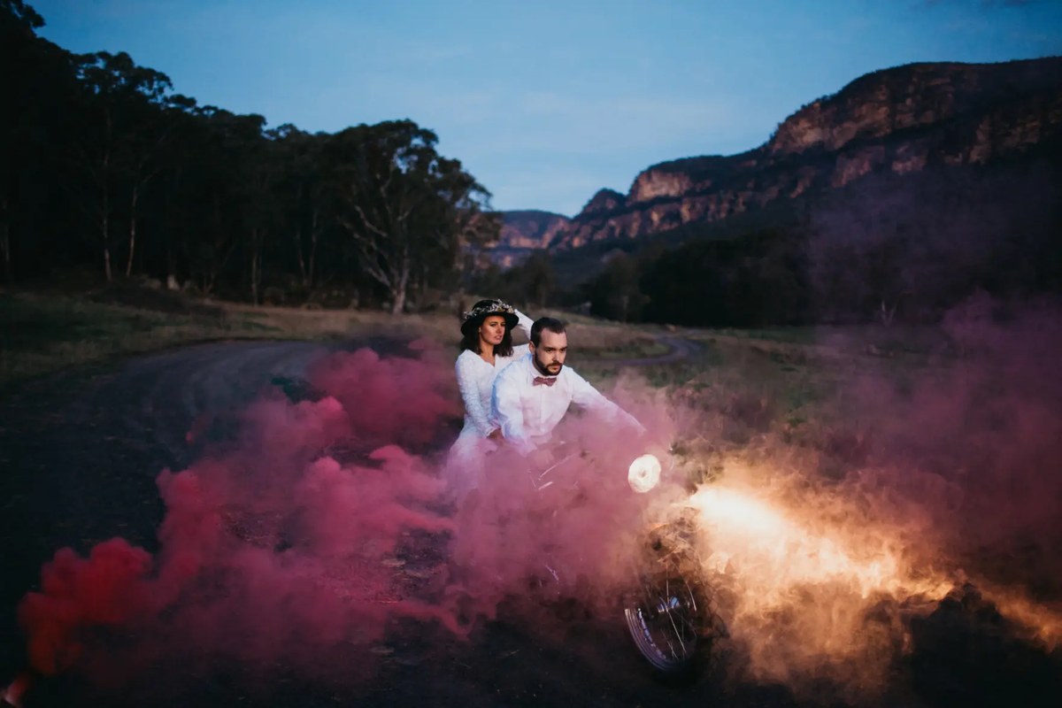 The bride and groom stand in colored smoke beneath a mountain. Photograph by documentary wedding photographer Joshua Mikhaiel.