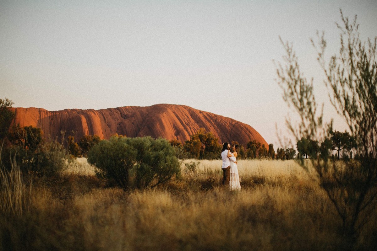 A couple stand together in tall grass with Uluru rising in the distance, photographed by documentary wedding photographer Joshua Mikhaiel.