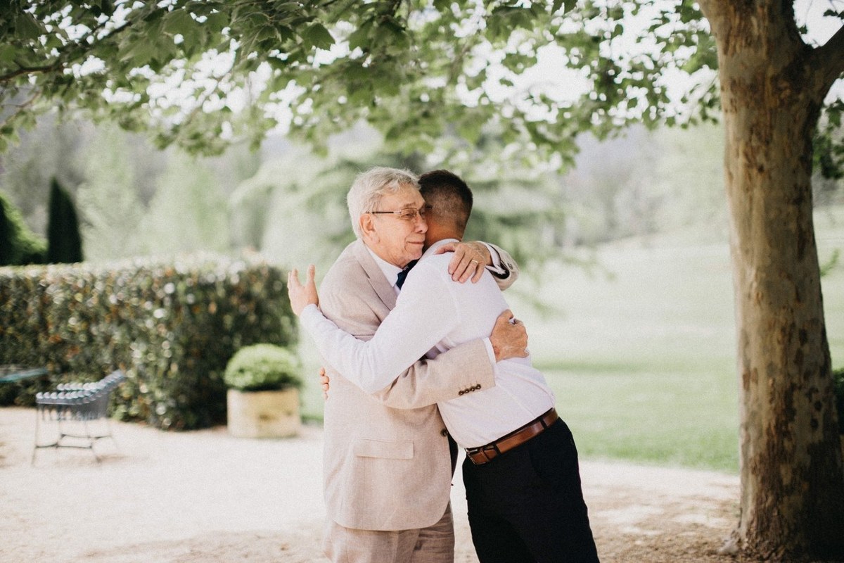 The groom embraces an older guest beneath the garden trees. Photograph by documentary wedding photographer Joshua Mikhaiel.