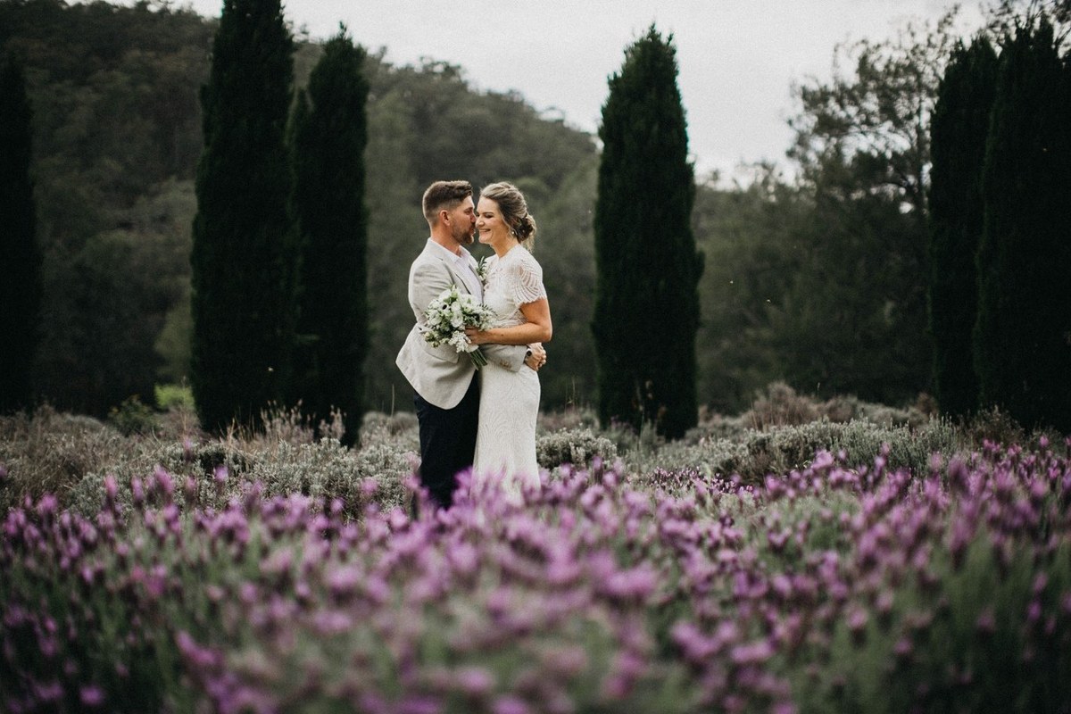 The bride and groom pose among a bed of purple flowers and tall trees. Photograph by documentary wedding photographer Joshua Mikhaiel.