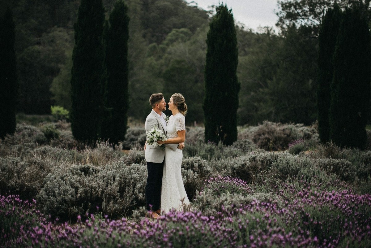 The bride and groom stand together among flowering shrubs and tall trees. Photograph by documentary wedding photographer Joshua Mikhaiel.