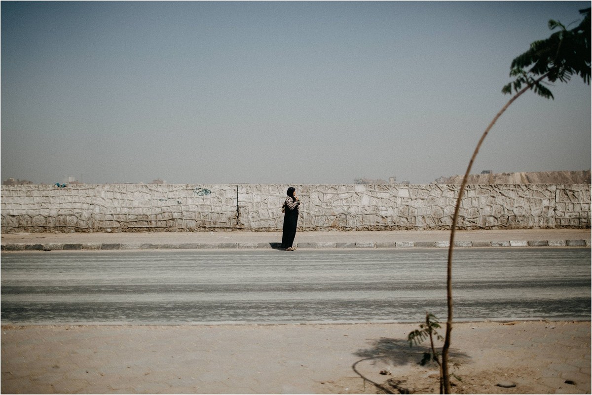 A lone person stands beside a broad riverbank with low buildings across the water in Egypt, photographed by documentary photographer Joshua Mikhaiel.