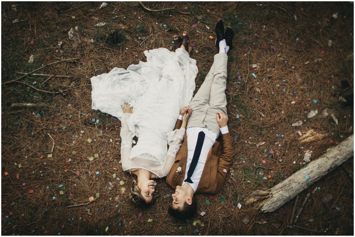 A bride and groom lie side by side on a leaf-strewn forest floor, photographed by documentary wedding photographer Joshua Mikhaiel.