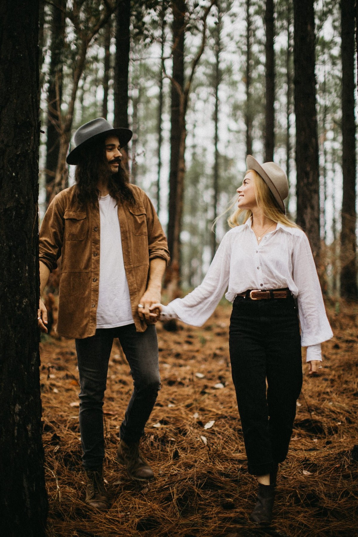 A couple hold hands between tall trees during an engagement session photographed by documentary wedding photographer Joshua Mikhaiel.