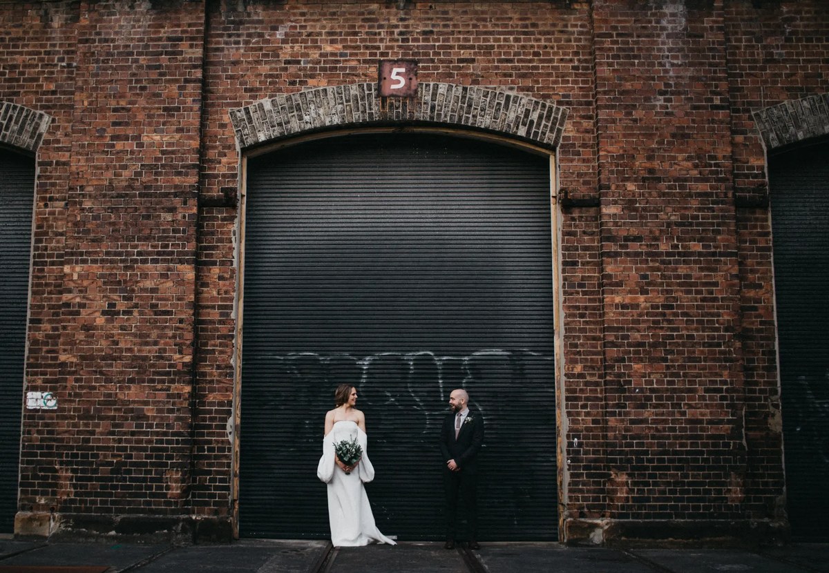 A couple stand before a large arched loading door in a brick building. Photograph by documentary wedding photographer Joshua Mikhaiel.