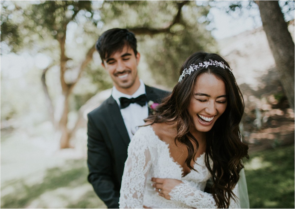 A bride laughs as the groom stands behind her among trees, photographed by documentary wedding photographer Joshua Mikhaiel.