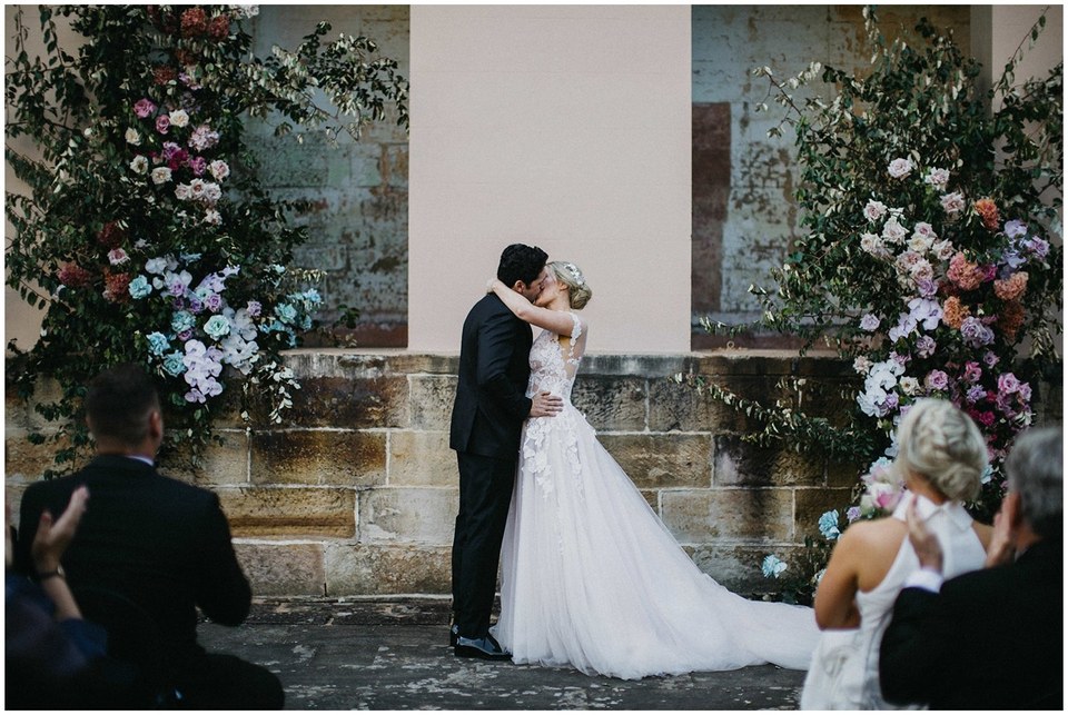 A bride and groom kiss between large floral arrangements during their outdoor ceremony, photographed by documentary wedding photographer Joshua Mikhaiel.