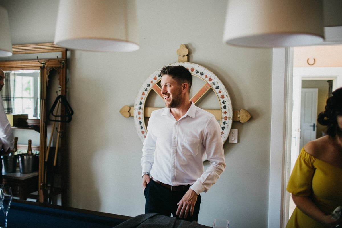 The groom stands in the room beside a round mirror as he gets ready. Photograph by documentary wedding photographer Joshua Mikhaiel.