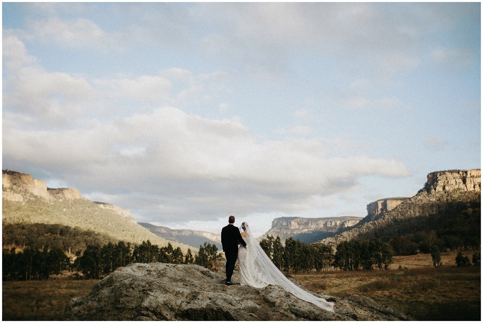 A bride and groom stand together beneath sandstone cliffs in a wide valley. Photograph by documentary wedding photographer Joshua Mikhaiel.
