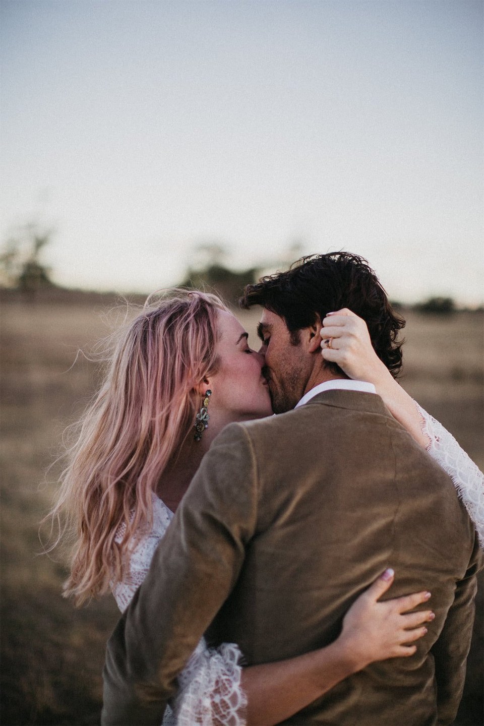A couple kiss in an open field as one holds a hat against the other’s head. Photograph by documentary wedding photographer Joshua Mikhaiel.